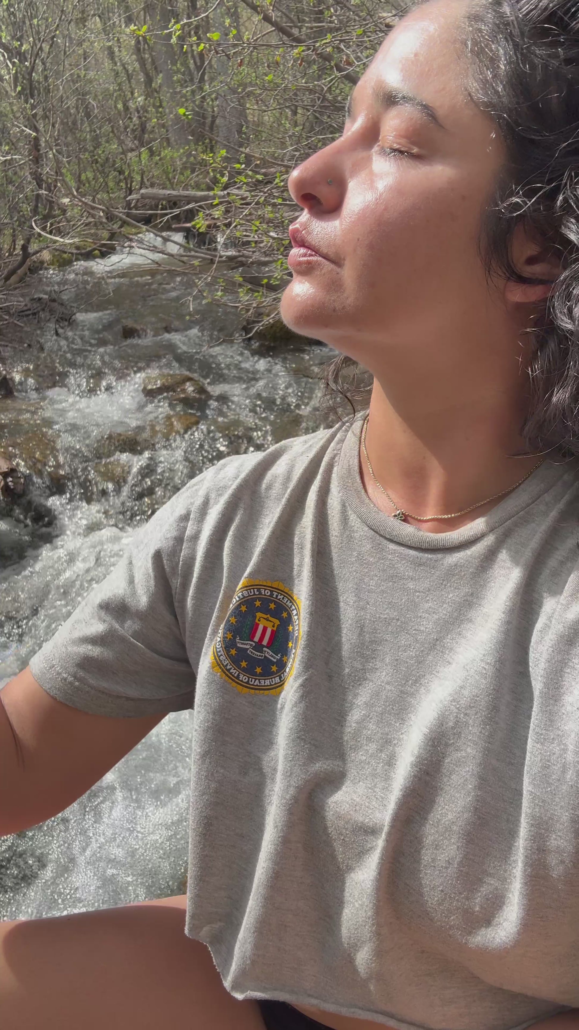 A woman closes her eyes to enjoy a refreshing mist of the•alambique organic microbiome facial toner while sitting by a sunlit mountain river stream.